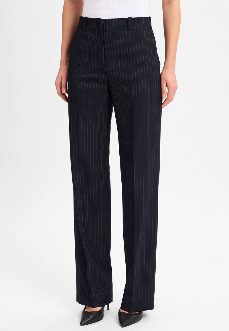 Vrouw in zwarte pinstripe wijdlopende broek met voorzakken en zwarte puntige hakken, staand tegen een witte achtergrond.