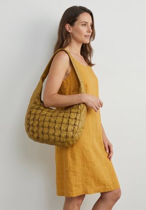 Woman wearing a sleeveless mustard dress carrying a large mustard shoulder bag with textured circular patterns.