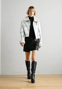 Chaqueta corta metálica plateada con cuello de piel, suéter negro de cuello alto, falda de cuero envolvente y botas negras hasta la rodilla. Textura suave, diseño ajustado.