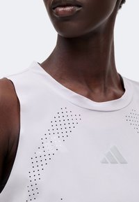 Gros plan d’une personne portant un haut blanc sans manches avec des motifs perforés et un logo Adidas discret sur la poitrine.
