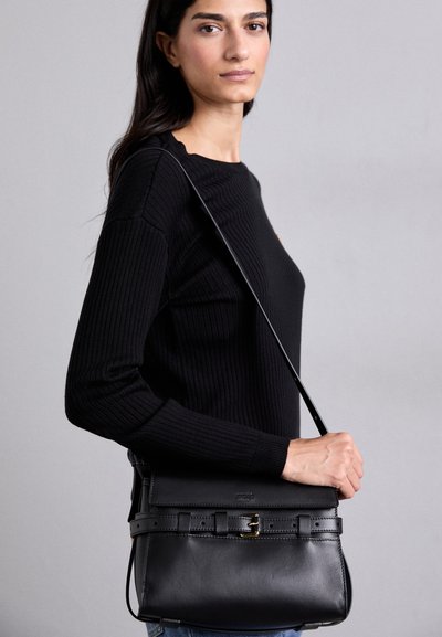 Mala de mão em couro preto com um design estruturado, apresentando um fecho de aba e um detalhe decorativo em forma de cinto. Usada como mala de lado com uma alça fina.