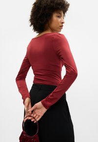 Dolga majica z dolgimi rokavi iz burgundskega blaga, z okroglim izrezom in gladko teksturo. Ujemajoča se s črnimi hlačami in burgundsko torbico.