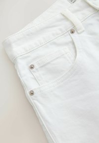 Blugi din denim alb, prim-plan, care prezintă buzunarul frontal, buzunarul mic pentru monede, bucla pentru curea și detalii metalice cu nituri pe un fundal deschis.