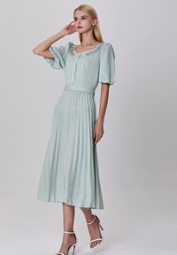 Robe vert menthe avec manches bouffantes, boutonnage sur le devant et jupe plissée. Présente un col orné de bijoux et une ceinture assortie. Portée avec des talons blancs.