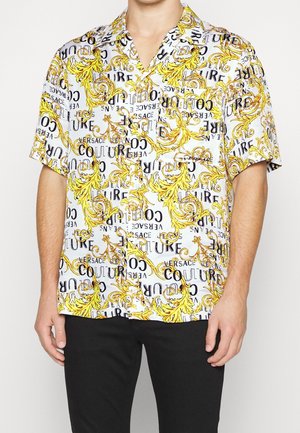 Weißes kurzärmeliges Hemd mit Knöpfen, gelbem barockem Muster und schwarzem "Versace Couture" Schriftzug, getragen von einer Person mit schwarzer Hose.