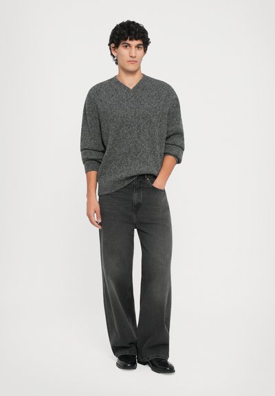 Gray, textured knit sweater with a V-neck. Paired with wide-leg black jeans. Model stands in black shoes, against a plain white background.