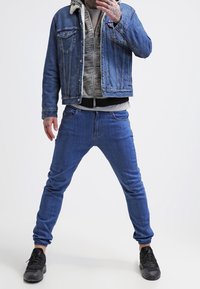 Uomo che indossa una giacca di jeans sopra un felpa grigia e jeans blu, stando in piedi con le gambe divaricate e una mano vicino al viso.