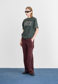 Zelené mramorované oversized tričko s logom "NIKE", skombinované s vínovými nohavicami s bielymi bočnými pásmi. Čierne slnečné okuliare a topánky dotvárajú vzhľad.