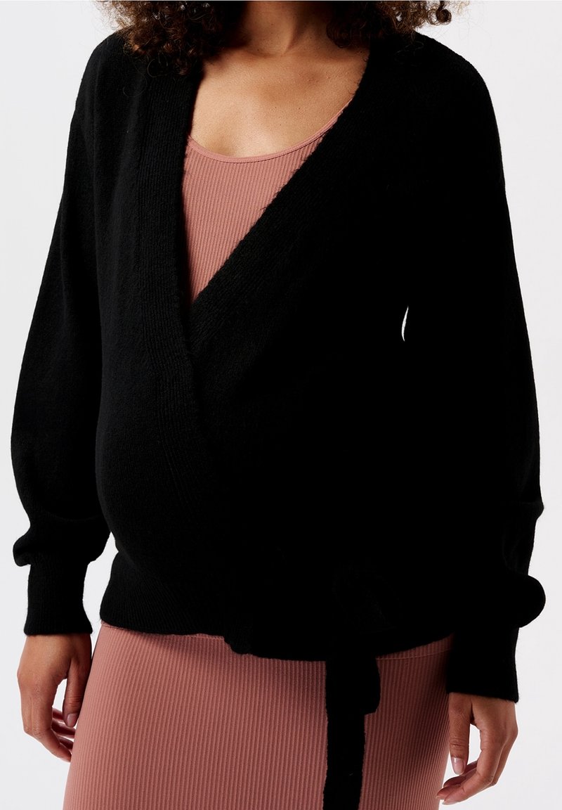 Cardigan noir croisé avec manches longues bouffantes, fabriqué en matériau doux. Présente une taille nouée et un col en V, associé à une robe rose côtelée.