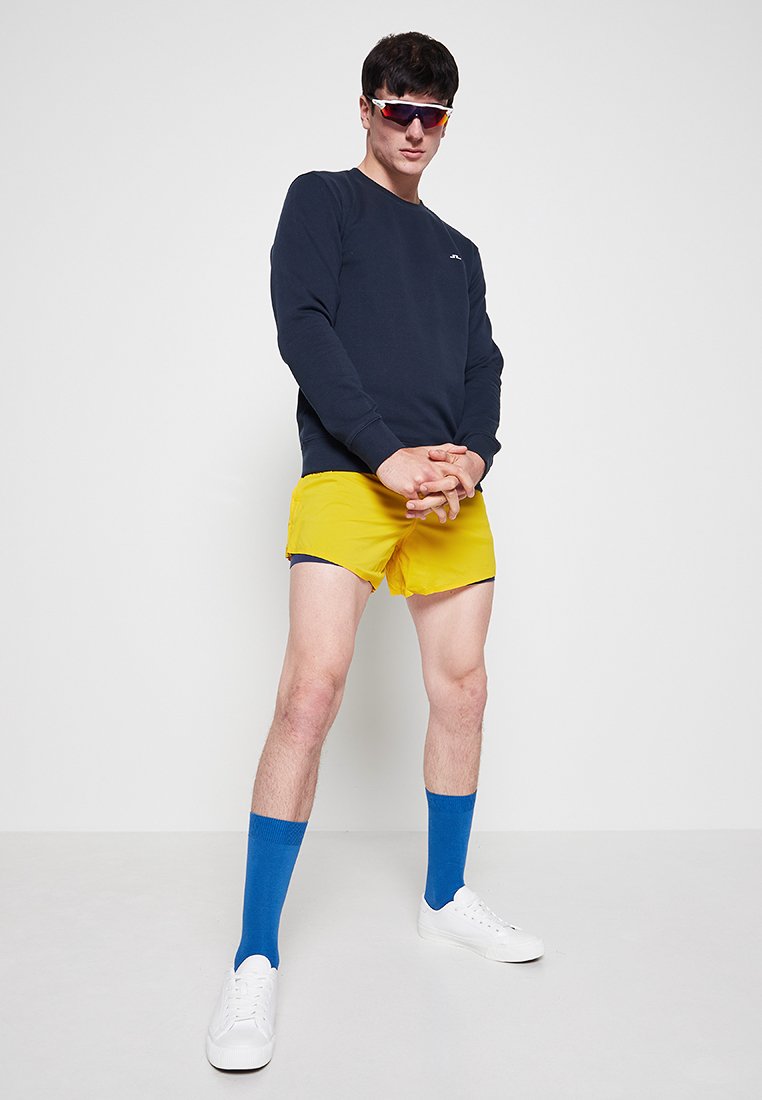 Marineblauer Sweatshirt, gelbe Shorts, blaue Kniesocken und weiße Sneaker. Modell posiert mit verschränkten Händen und trägt Sonnenbrille. Einfarbiger Hintergrund.