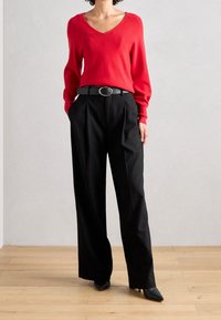 Czerwony sweter w kształcie litery V, miękka dzianina; czarne spodnie z wysokim stanem i plisami z przodu; eleganckie czarne szpilki; minimalistyczne otoczenie z drewnianą podłogą.