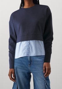 Sweat-shirt bleu marine avec un design raccourci et des manches longues, superposé sur une chemise à rayures bleu clair et blanc avec des boutons à l'ourlet.