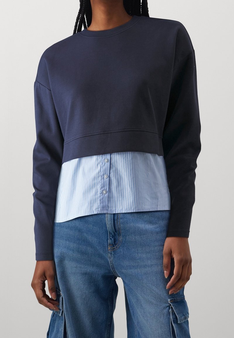 Sweat-shirt bleu marine avec un design raccourci et des manches longues, superposé sur une chemise à rayures bleu clair et blanc avec des boutons à l'ourlet.