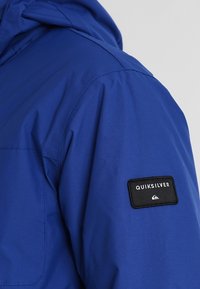 Modrá bunda s kapucí, s hladkým povrchem a černou nášivkou loga Quiksilver na levém rukávu. Odolný materiál.