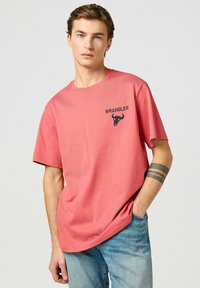 Camiseta de algodón color coral con cuello redondo y mangas cortas. Presenta un logo negro de "WRANGLER" y un gráfico de un toro en el lado izquierdo del pecho.
