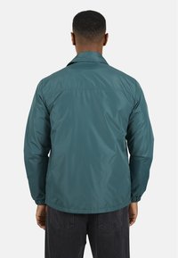 Giacca leggera color turchese con texture liscia, design semplice e colletto, dotata di polsini e orlo elasticizzati. Vista posteriore mostrata.