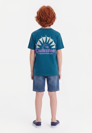 Barn med krøllet rødt hår iført en teal Quiksilver t-shirt, denimshorts og marineblå sneakers, stående med ryggen til på en hvid baggrund.