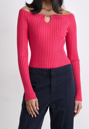 Vrouw draagt een aangesloten, geribd, felroze off-shoulder top met een kleine voorste sluiting en donkerblauwe hoge taille broek.