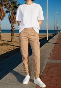 Personne portant un t-shirt blanc, un pantalon cargo beige et des baskets blanches, debout sur une promenade en bord de mer avec des palmiers et l'océan en arrière-plan.