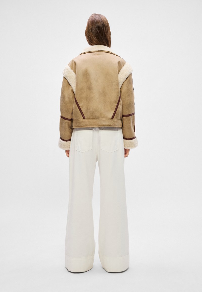 Brauner Lammfelljacke mit weißem Pelzkragen und -manschetten, die bordeauxrote Akzente und ein verkürztes Design aufweist. Kombiniert mit weißen weit geschnittenen Hosen.