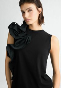 Top nero senza maniche con un’accento di tessuto arricciato e strutturato su una spalla. Design liscia e aderente con scollatura rotonda.