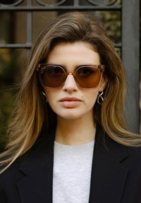 Hnědé oversized sluneční brýle s tlustými obroučkami, jemným květinovým vzorem a zlatými akcenty. Modelka má na sobě šedý top a černý blejzr.