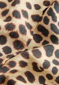Tissu beige avec des taches irrégulières de léopard noir et marron, montrant des plis et des lignes de couture visibles.