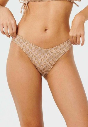 Kobieta ubrana w beżowy wzorzysty dół i górę bikini, regulująca boki dołu bikini na jednolitym jasnym tle.