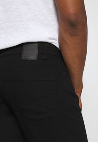 Jeans de mezclilla negra con una textura suave, que cuentan con un bolsillo trasero y una etiqueta de cuero en la cinturilla. El diseño es ajustado.
