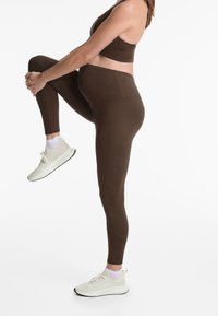 Bruna stretchleggings med hög midja, i kombination med en matchande brun sportbh och ljusa träningsskor.