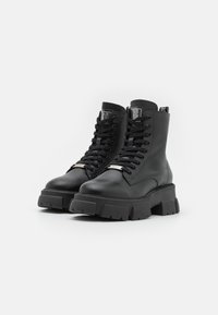 Bottes de combat en cuir noir avec semelles épaisses à crampons, lacets à l'avant et languettes de tirage marquées sur fond blanc.