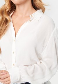 Blusa bianca con texture e una morbida caduta, colletto bottonato e ampie maniche a cuffia. Presenta sottili dettagli in oro sui bottoni.
