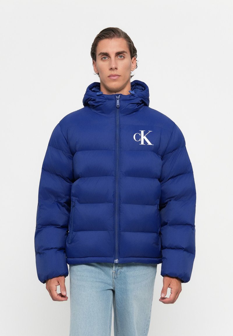 Mand iført en blå pufferjakke med hætte og hvidt "cK" logo på brystet, parret med lyseblå jeans, stående mod en ensfarvet baggrund.