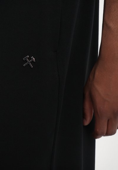 Schwarze Hose mit glattem Textur, die ein kleines metallisches Logo auf der Tasche aufweist. Der Stoff wirkt leicht und maßgeschneidert.
