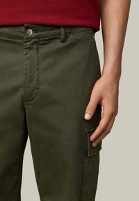 Olijfgroene cargobroek met knoopsluiting, zijzak en een gladde textuur. Draagt een rood shirt, hand rustend op de stof.