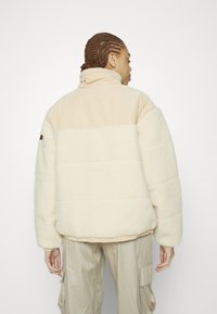 Krämfärgad vadderad jacka med en mjuk, texturerad fleece nederdel och en slät tyg övre del, med hög krage och elastiska muddar.