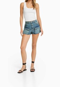 Nainen, jolla on yllään valkoinen hihaton paita, korkeavyötäröiset farkkushortsit suurilla etutaskuilla ja mustat nauhalliset litteät sandaalit, seisoo valkoista taustaa vasten.