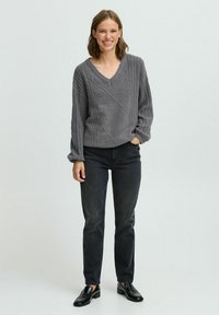 Grauer Kabelstrickpullover mit V-Ausschnitt und lockeren Ärmeln, kombiniert mit dunklen Jeans und schwarzen Schuhen, vor einem schlichten Hintergrund.