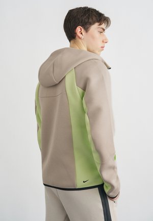 Giovane di profilo che indossa una giacca con cappuccio beige e verde chiaro con il logo Nike nero sulla parte bassa della schiena, pantaloni chiari con striscia nera.