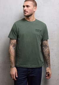 Zelené bavlněné tričko s kulatým výstřihem a krátkými rukávy. Logo "Tough Trails" tištěné tmavě zelenou barvou na levé straně hrudi.