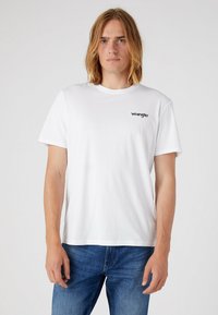 Camiseta de algodón blanca con mangas cortas y cuello redondo, con un pequeño logo negro de "Wrangler" en el lado izquierdo del pecho.
