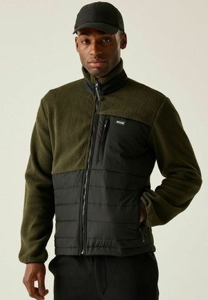 Mężczyzna w zielonej i czarnej polarowej kurtce oraz czapce z daszkiem, stojący z jedną ręką w kieszeni kurtki na jednolitym tle.