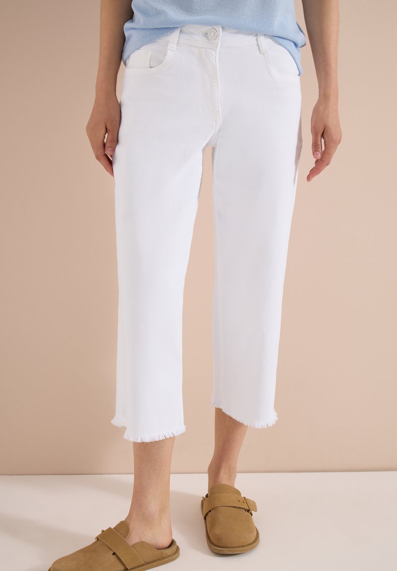 Mujer con vaqueros blancos cortos con bajos deshilachados y zapatos sin cordones color beige, de pie frente a un fondo beige.