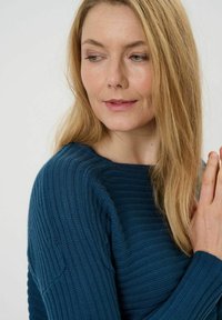 Strickpullover in Teal mit gerippter Textur, breitem Ausschnitt und langen Ärmeln. Das Modell hat blonde Haare und gefaltete Hände.