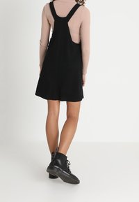 Czarna bawełniana sukienka ogrodniczka z dopasowanym górnym krojem i rozkloszowaną spódnicą. Noszona na beżowej bluzce z długim rękawem, zestawiona z czarnymi botkami na sznurowadła.