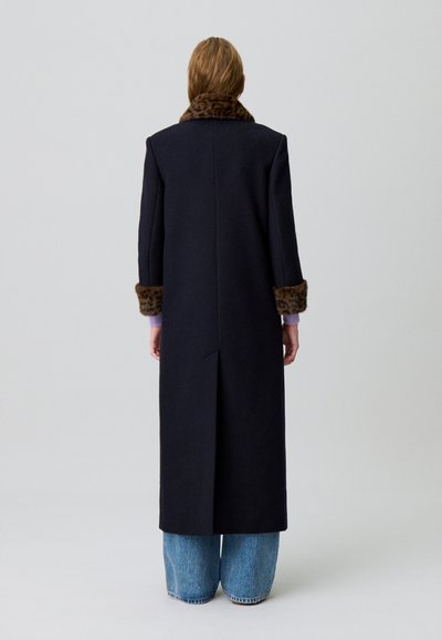 Abrigo de lana navy con cuello y puños de piel sintética marrón, silueta recta, hasta la rodilla, detalle de ventilación en la parte posterior, llevado sobre vaqueros de pierna ancha.