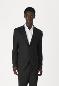 Blazer à carreaux noirs en tissu texturé, doté d'un design à deux boutons, de revers en pointe et de poches latérales, associé à une chemise blanche.