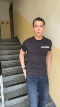 Νέος άνδρας με μαύρο t-shirt "Replay" και σκισμένο τζην στέκεται σε σκάλες σε έναν διάδρομο κτιρίου βαμμένο σε κίτρινο-μπεζ χρώμα.