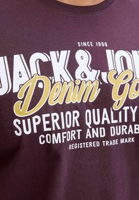Burgundi puuvillane T-särk, millel on julge valge ja kollane tekst: "JACK & JONES Denim Go" ja "Üksikasjalik Kvaliteet, Mugavus ja Vastupidavus."