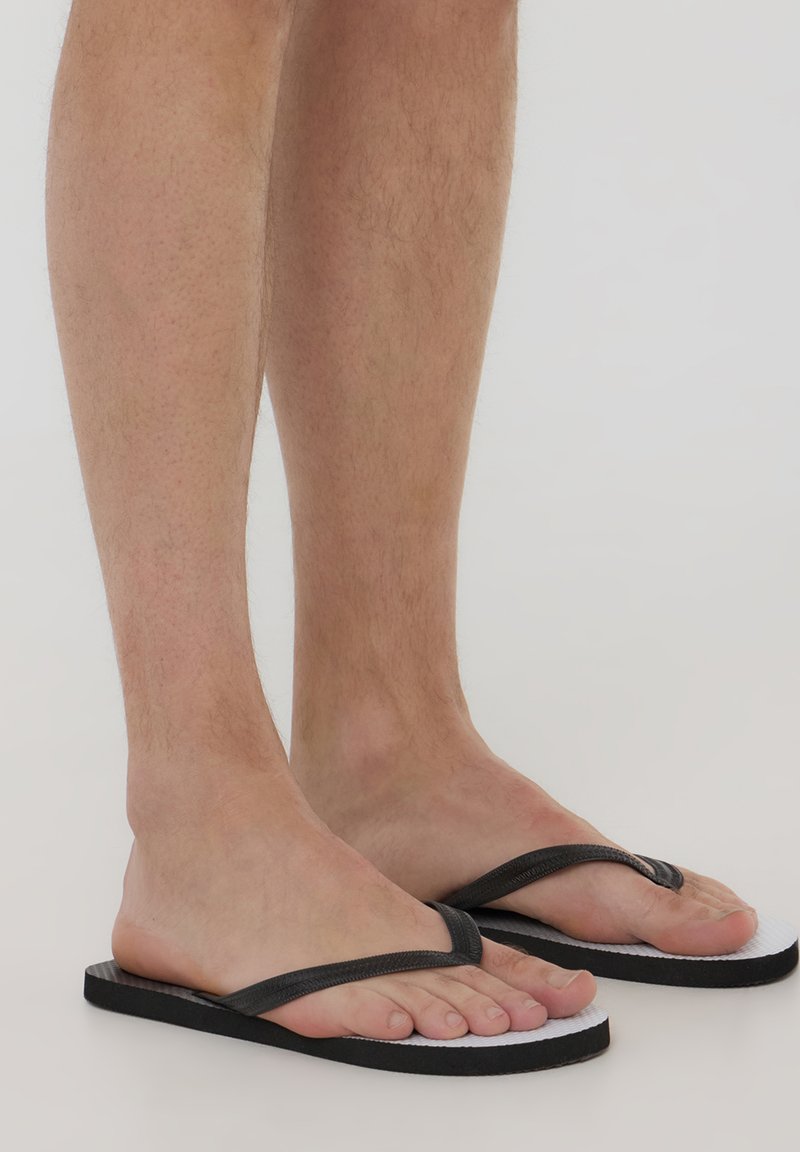 Infradito nere in gomma con una fascia testurizzata e suola bianca, caratterizzate da un design semplice e profilo piatta, indossate su piedi nudi.